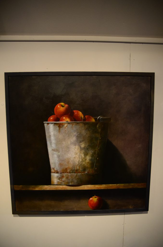 Bilden visar en målning av färgstarka äpplen i en plåthink.